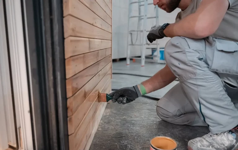 Eine Person renoviert eine Holzwand mit Pinsel, Handschuhen und Arbeitskleidung.