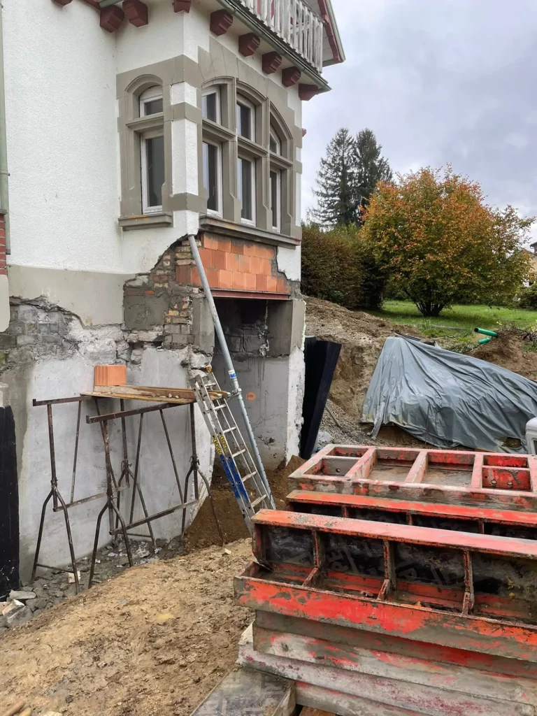 Unter einer Hauswand wird für Betonarbeiten ein Loch gebohrt, das durch ein Gerüst abgestützt wird.